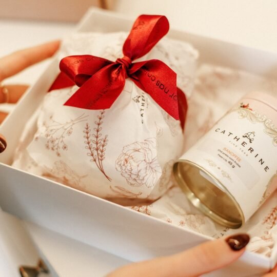 Uma "caixa de afeto" Catherine Fine Teas sendo aberta, revelando a icônica Pink Tin e louças, materializando o ritual sofisticado de presentear com chá.