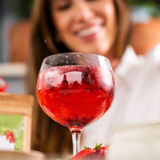 Close em taça balão suada, repleta de Iced Tea de frutas vermelhas vibrante (Red Velvet ou Strawberry Fresh) com muito gelo. Ao fundo, desfocada, a fundadora Ana Carolina Delai sorri, com uma embalagem da linha Iced Tea visível.