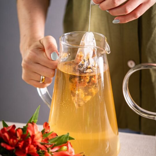 Detalhe do sachê artesanal de 14g da Catherine Fine Teas sendo imerso em jarra de vidro. O sachê contém ingredientes inteiros (frutas e folhas) que se expandem na água, ideal para preparar 1 litro de chá gelado.
