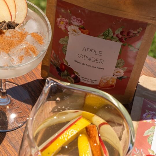Embalagem do Blend Apple Ginger - Iced Tea de Frutas e Flores da Catherine Fine Teas, apresentada em uma mesa ensolarada ao lado de uma taça de chá gelado com gelo, maçã e especiarias, e uma jarra com a infusão de frutas frescas.