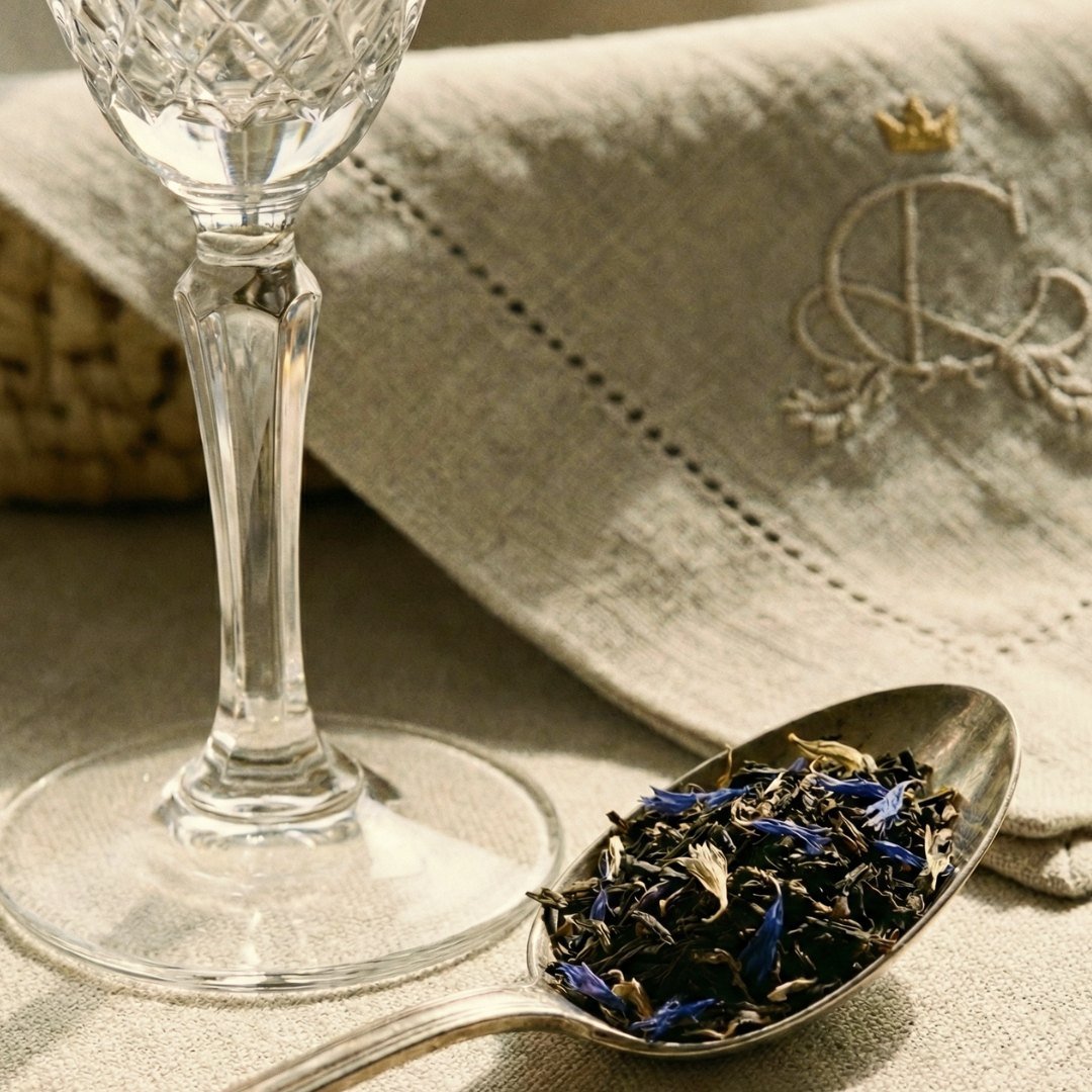 Detalhe em macrofotografia revelando a textura de folhas inteiras de chá Earl Grey Blue Flower da Catherine Fine Teas de alta qualidade. O contraste visual é feito com o brilho da base de uma taça de cristal lapidado e a suavidade da asa de uma xícara de porcelana, ressaltando a excelência dos materiais.